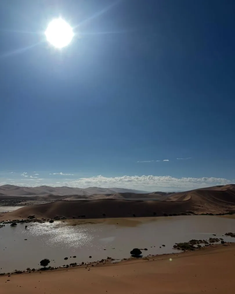 The sun bursting over dunes above a thin sheet of water on the sand