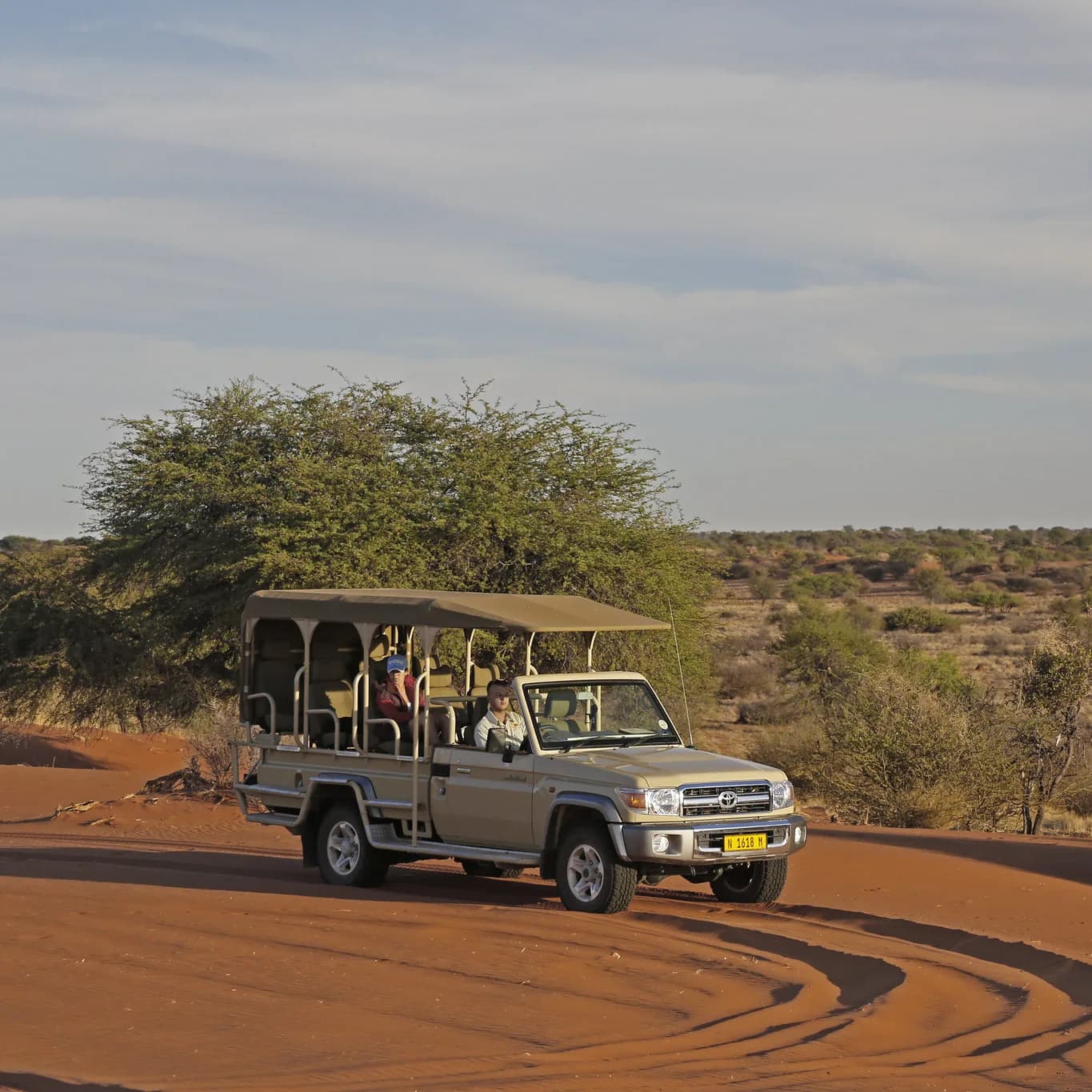 Open safari vehicle with two guests driving over red sand at golden hour