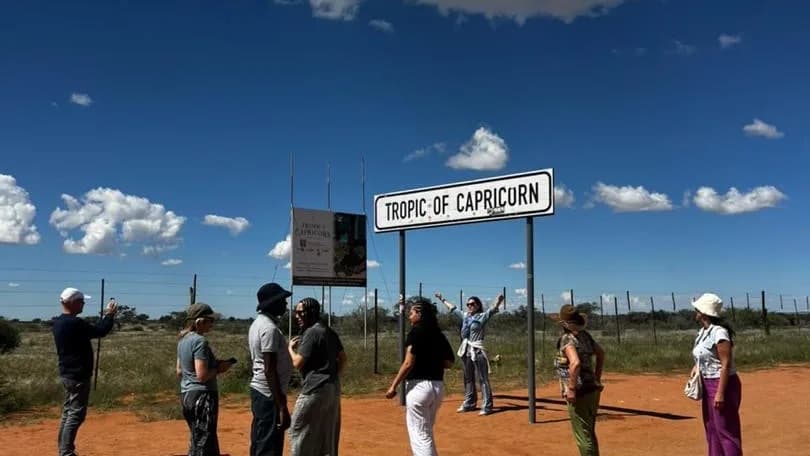 Tour guests gathered around the Tropic of Capricorn roadside sign on red desert soil