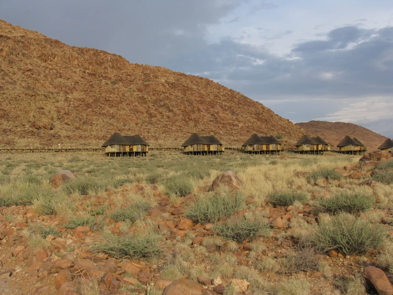 Row of thatched chalets on stilts below a rocky red hillside under a clear sky