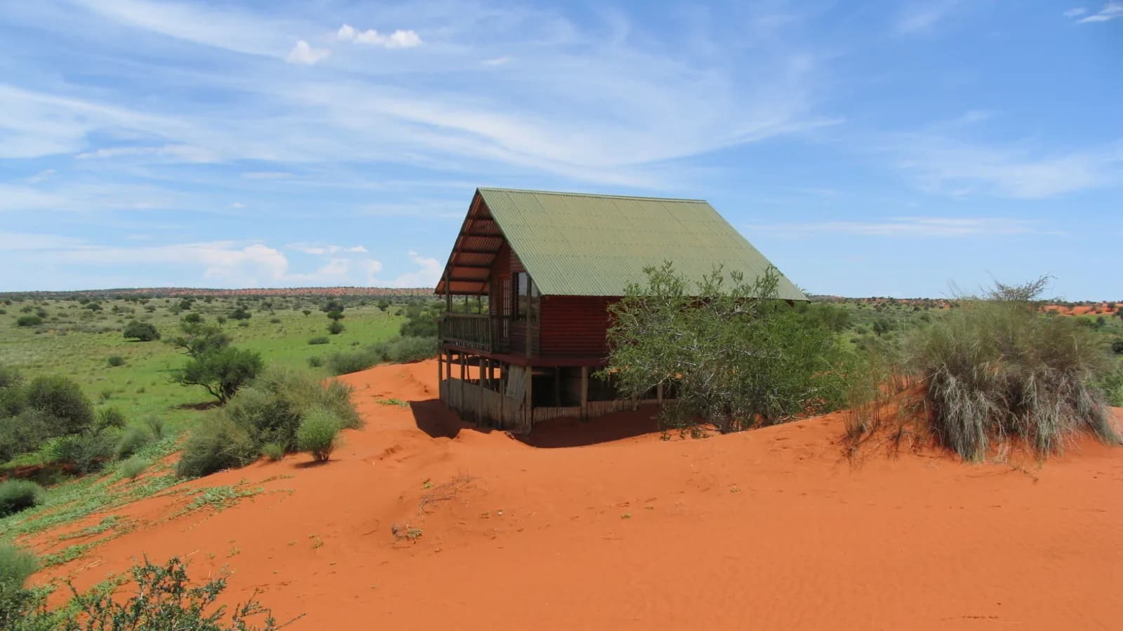 Timber lodge cabin with a green roof standing on red Kalahari sand under a clear blue sky