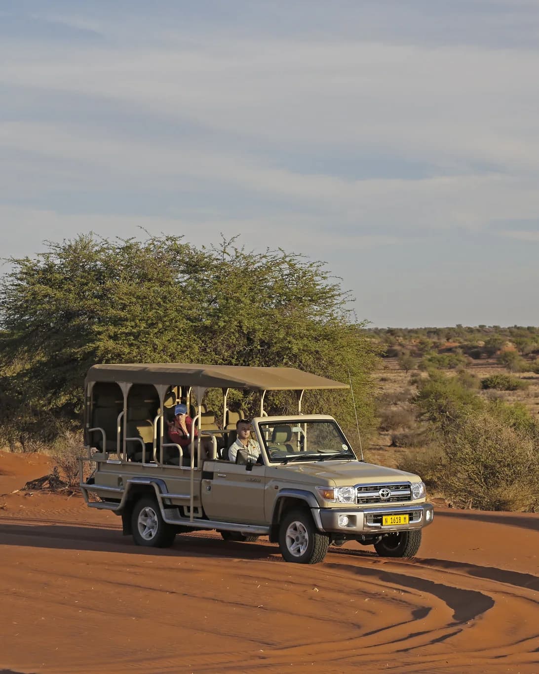 Open safari vehicle with two guests driving over red sand at golden hour
