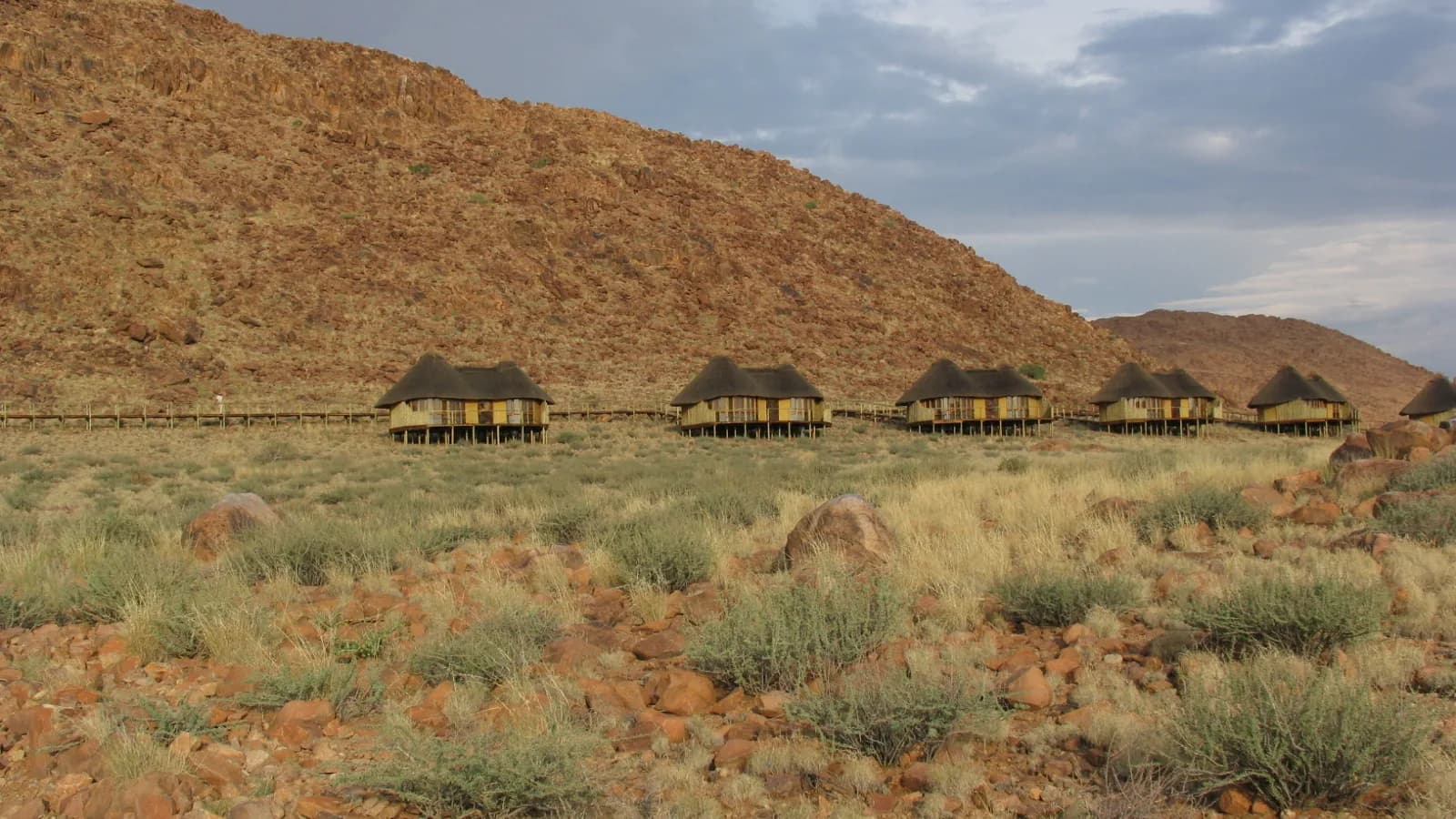 Row of thatched chalets on stilts along a rocky red hillside, seen from below at midday